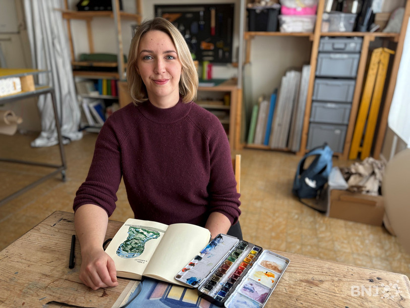 Une femme assise au bureau d'artiste, dans un atelier, avec un cahier et une boite de peinture à l'aquarelle devant elle.