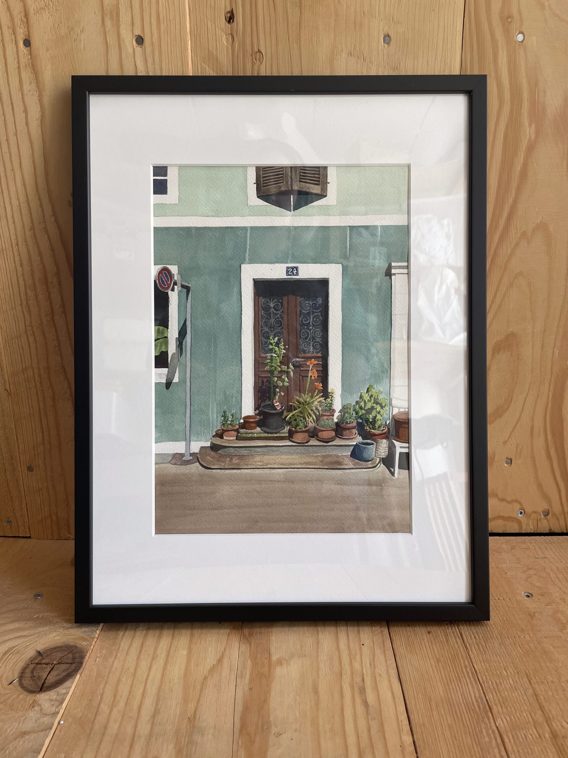 Peinture à l'aquarelle d'une porte en bois à La Chaux-de-Fonds, entre des murs vertes, avec des plants dans l'avant plan. Encadré en noir.