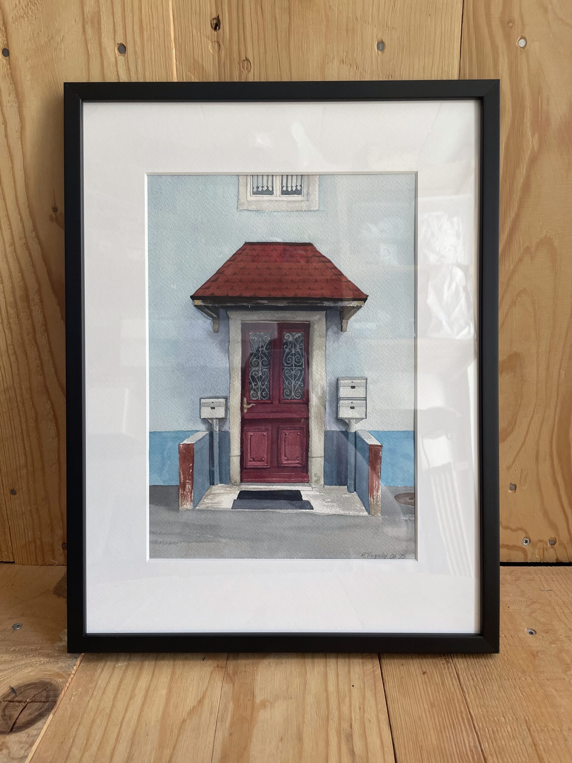 Une peinture à l'aquarelle d'une porte rouge et une porche rouge, contre des murs bleu. Le tableau est encadré en noir.