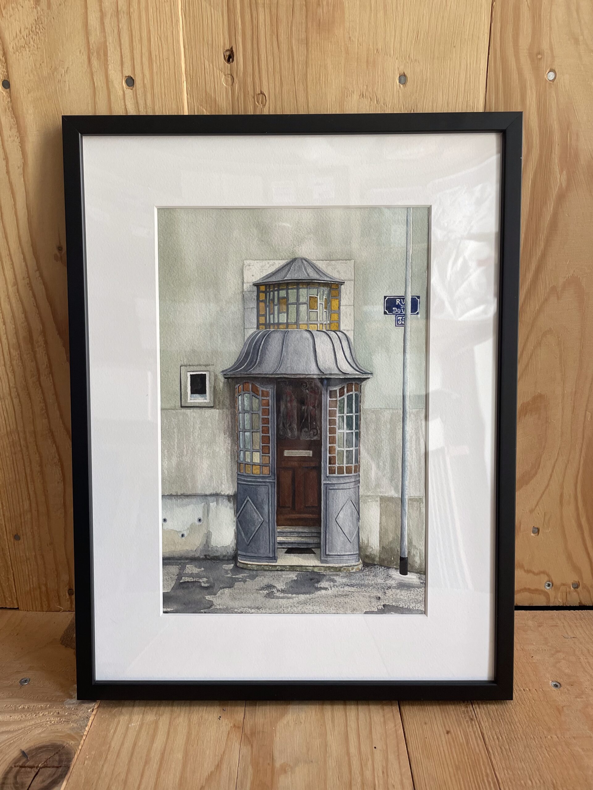 Peinture à l'aquarelle d'une porte en bois à La Chaux-de-Fonds, entre des murs blanches, avec une porche dans le style art nouveau. Encadré en noir.